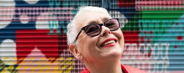 older woman in red leather jacket and sunglasses stands smiling in front of a colourful mural with her chin tilted upwards towards the sky.
