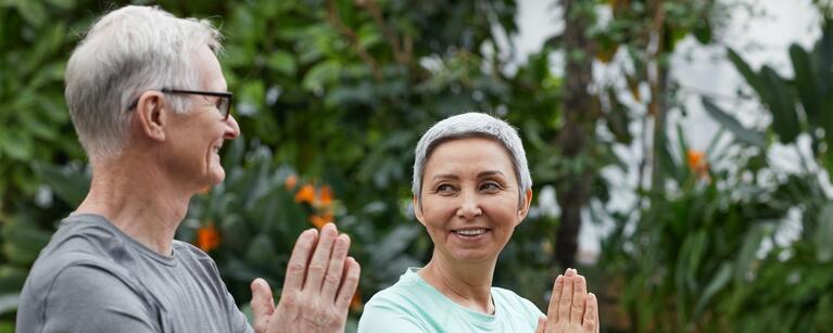 older man and woman stand outside in a yoga pose smiling towards each other.