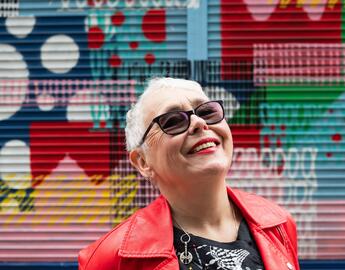 older woman in red leather coat and sunglasses smiling with chin tilted up, face towards the sky in front of a colourful mural.