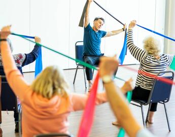 A group of people are sitting on chairs, facing a physical activity instructor. The participants and the instructor are holding a resistance band overhead. 
