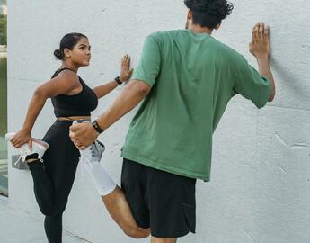 A man and woman looking towards one another as they are holding the wall with 1 hand, doing a quadricep stretch